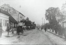 Zdjęcie czarno-białe  ukazujące fragment  ulicy Rynek Zygmunta Augusta, po prawej stronie widoczna słabo część augustowskiej cerkwi a na chodniku stoi grupa mężczyzn, natomiast po lewej stronie po ulicy jedzie dorożka oraz widać w oddali spacerujących ludzi.