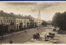 Zdjęcie czarno- białe ukazujące pierzeję południową ulica Rynek Zygmunta Augusta, na pierwszym planie po prawej stronie jest kilka drewnianych wozów konnych czyli furmanek - na dwóch są ludzie a po lewej są augustowskie kamieniczki- w oddali widoczny ratusz oraz spacerujący ludzie.