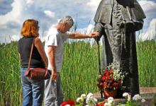 Pomnik św. Jana Pawła II w Studzienicznej, przed którym stoi mnóstwo biało- czerwonych kwiatów oraz dwa znicze, po lewej stronie zdjęcia jest kobieta i mężczyzna. Mężczyzna położył swoją dłoń na pomniku  fot. J. Koniecko