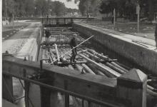 Zdjęcie czarno-białe przedstawiające spław drewna na Kanale Augustowskim w trakcie śluzowania na Śluzie Augustów w 1930 roku, widoczny na pierwszym planie flisak na tratwie.