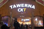 Zdjęcie przedstawia Tawernę CKT z zewnątrz, widoczne duże drewniane wrota nad którymi jest podświetlony napis Tawerna CKT. Na zdjęciu widać również bardzo dużo osób. W tle widoczne podwieszane u góry telewizory.